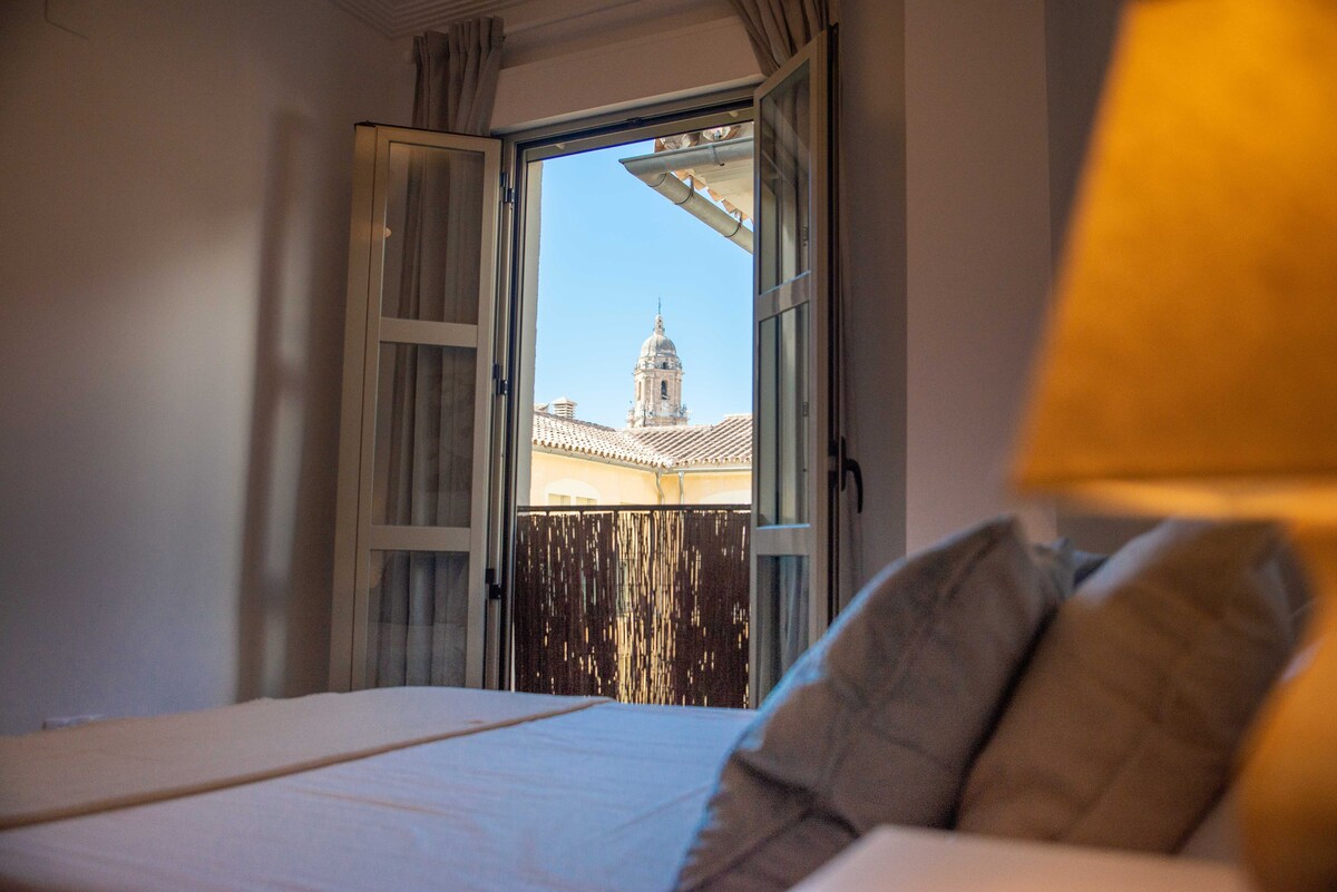 A view from a bedroom window is framed by two open doors, revealing a clear blue sky and a distant tower. The room features a spacious bed with light-colored bedding and a bedside lamp emitting a soft glow.