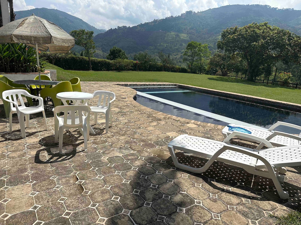 An outdoor area features a large swimming pool surrounded by lounge chairs under an umbrella. A dining table with colorful chairs is visible, set against a backdrop of lush hills and clear skies. The spacious patio is made of patterned stone, creating an inviting atmosphere.