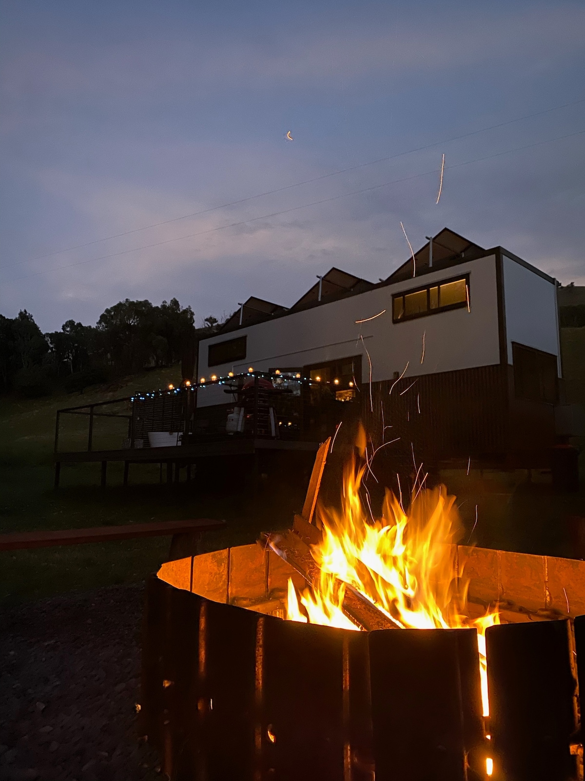 A fire pit is depicted in the foreground, featuring glowing flames and flying sparks. In the background, a modern tiny house is visible with an outdoor deck illuminated by soft lighting. The evening sky casts a serene blue hue over the scene.