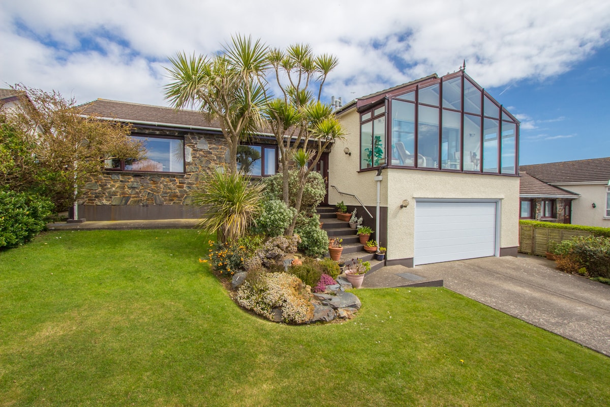 A charming exterior of a stone and rendered house is showcased, featuring a sloped lawn with colorful flower beds. A glass conservatory extends from the building, while palm trees add a tropical touch. A driveway is visible, leading to a garage on the lower level.