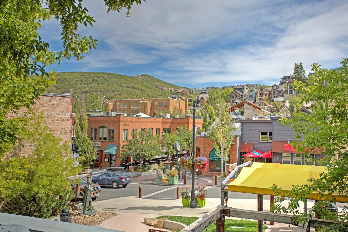 A view of a vibrant streetscape in Old Town features a mix of historic and modern buildings. Trees line the street, and colorful outdoor seating areas are visible. The backdrop includes rolling green hills under a partly cloudy sky.