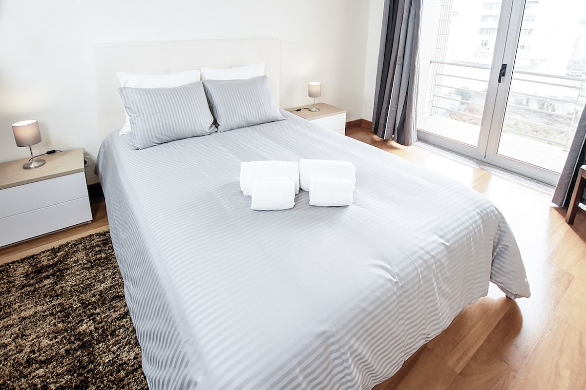 A spacious bedroom features a large bed dressed in light grey striped linens. Two bedside tables with lamps are positioned on either side. Natural light filters in through a nearby sliding glass door, leading to a balcony.