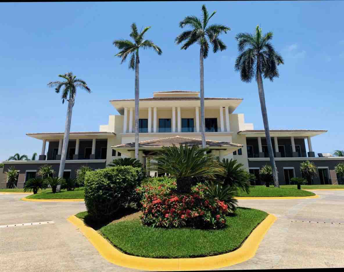 A modern multi-story building is framed by palm trees and lush greenery. A circular driveway leads to the entrance, which features a large overhang. Colorful flowers are situated in garden areas around the property, enhancing the inviting facade.