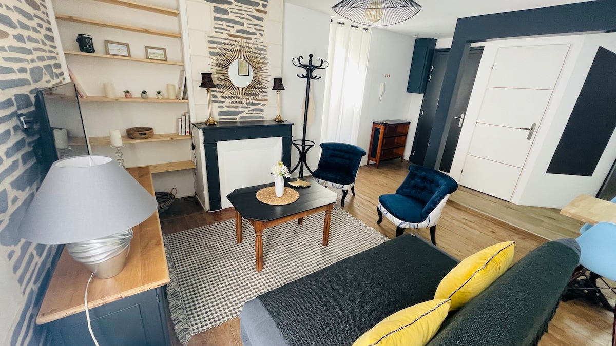 A living area is presented with a mix of modern and rustic elements. A dark wooden table is centered, surrounded by two upholstered blue chairs. A cozy sofa with grey upholstery sits adjacent. Decorative shelves and a wall mirror enhance the inviting ambiance.