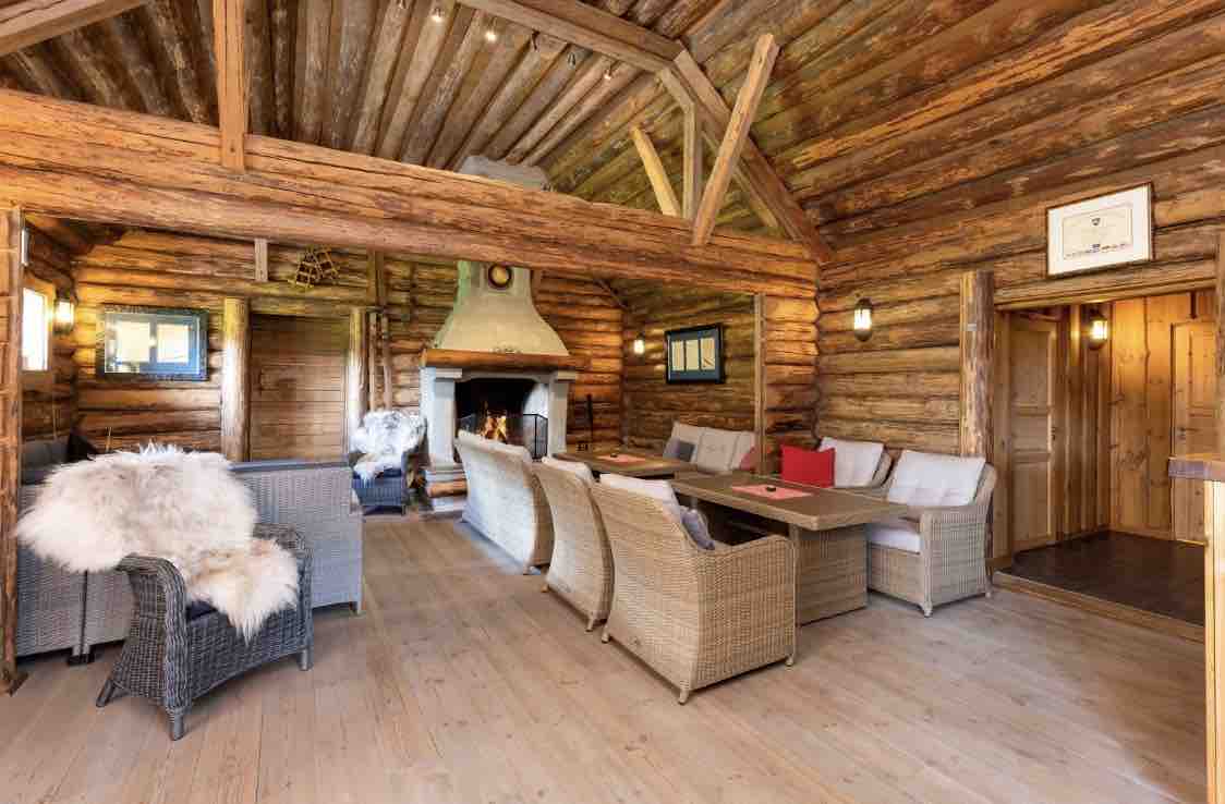A spacious living area features rustic wooden beams and a stacked log wall. Comfortable seating is arranged around a central table, with a cozy fireplace providing warmth. Natural light filters in through windows, enhancing the inviting atmosphere of the space.