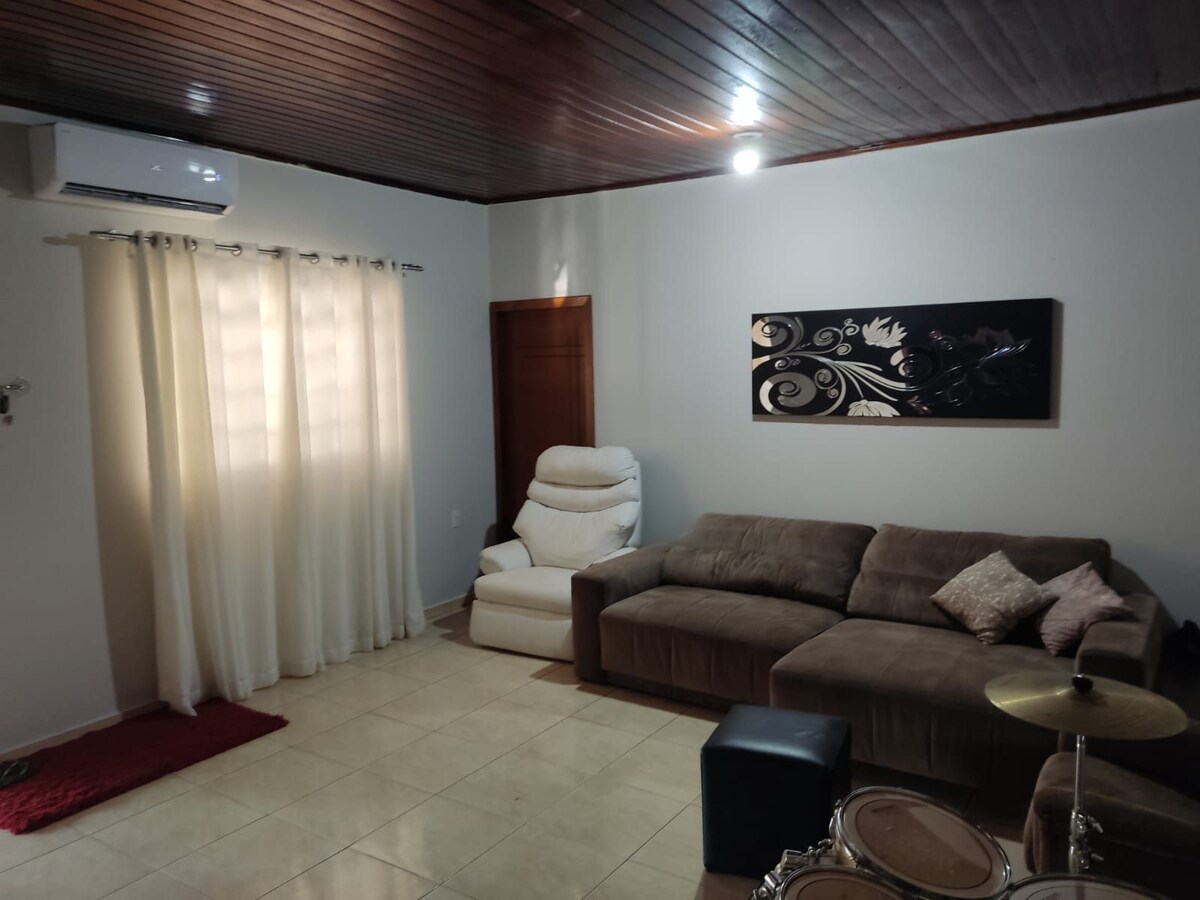 A comfortable living room is presented with a spacious sectional sofa, complemented by a reclining chair. Sunlight filters through sheer curtains, illuminating the light-colored tiled floor. A decorative wall art piece adds a touch of character to the room.