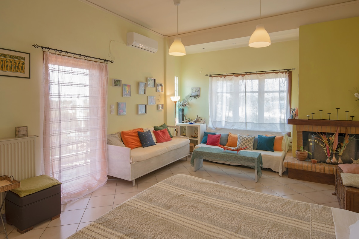 A spacious living area features light-colored walls and elegant lighting. Comfortable seating options are arranged around a central area, with soft cushions in shades of orange and teal. Natural light fills the space through large windows, and a cozy fireplace stands in one corner.