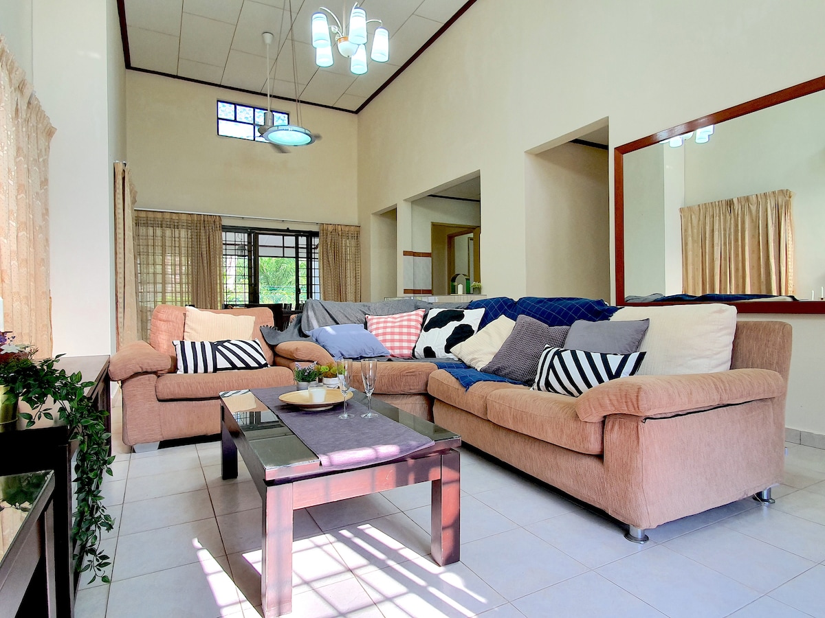 A spacious living area features a large sectional sofa arranged around a glass coffee table. Natural light enters through the windows, illuminating the room. Soft curtains offer privacy, while a mirror reflects the comfortable atmosphere. The space is designed for relaxation and socializing.