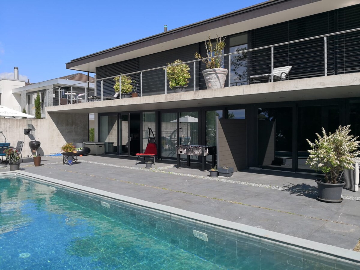 A modern two-story villa is depicted with large glass doors and a spacious terrace overlooking a swimming pool. Potted plants are positioned along the railing. Lounge chairs are set near the water, enhancing the outdoor living space.