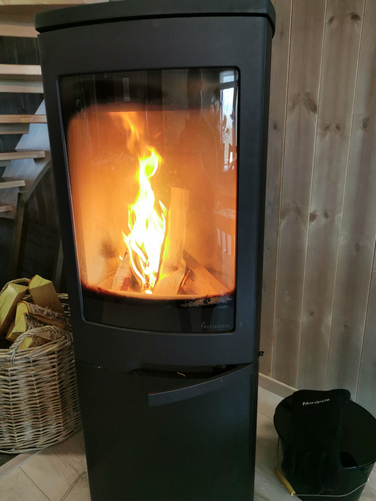 A modern wood-burning stove is featured, with flames dancing inside a transparent glass chamber. The surrounding area includes a woven basket for firewood and a pair of gloves positioned nearby, set against a backdrop of wooden wall panels.