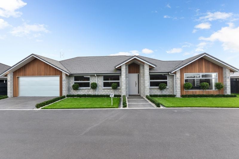 A modern, single-story house is showcased with a blend of stone and wood accents. Neatly trimmed hedges line the walkway leading to the front entrance. A spacious driveway and an attached garage are visible, complemented by a green lawn under a bright blue sky.