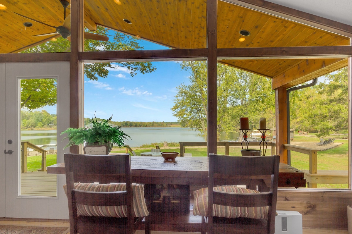 A dining area features a wooden table surrounded by upholstered chairs, with large glass windows framing a serene view of Lukens Lake. Natural light illuminates the space, and the ceiling showcases warm wooden beams. Lush greenery is visible outside, enhancing the tranquil setting.