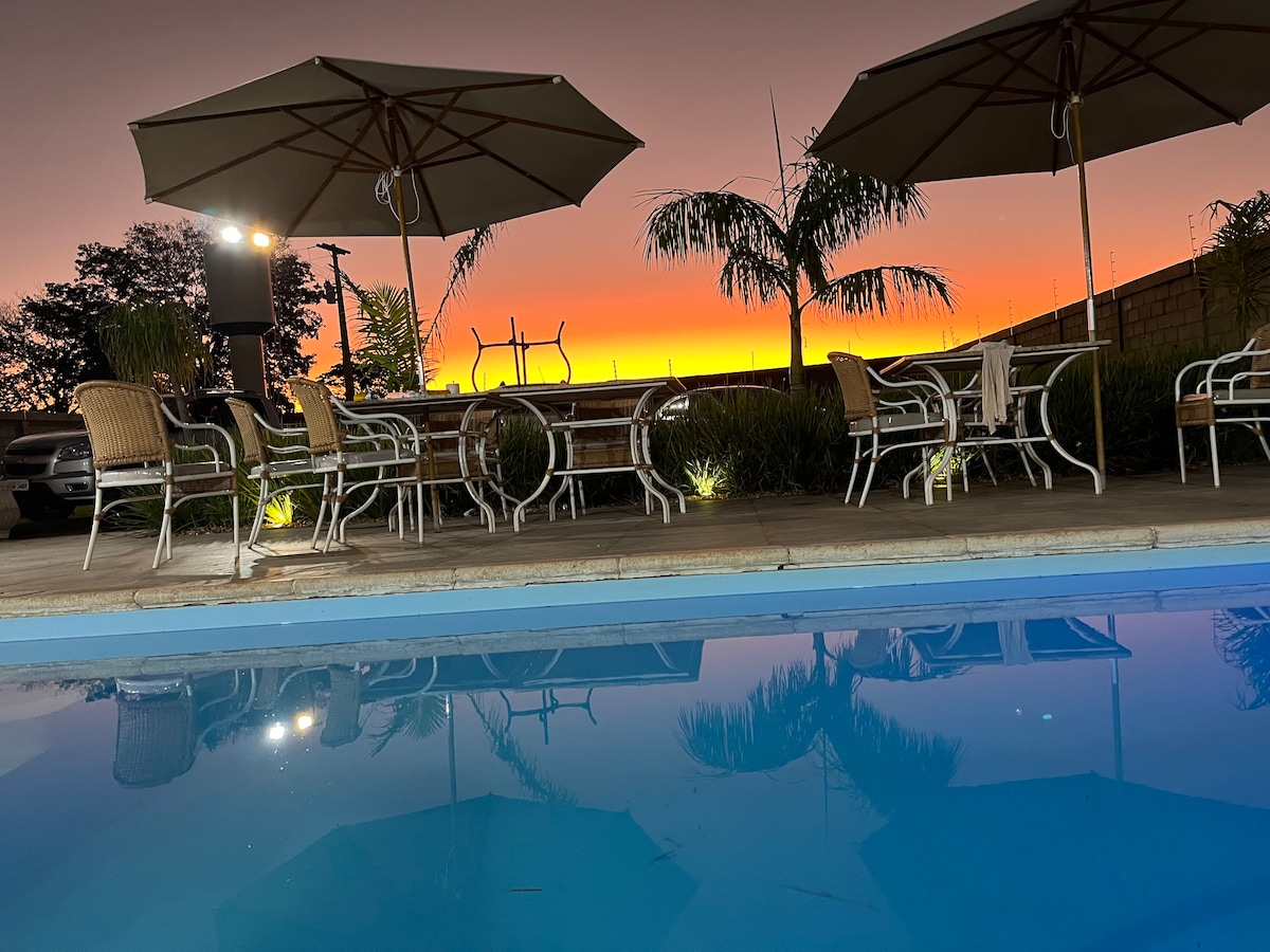 Beach loungers and dining chairs are arranged under large umbrellas beside a swimming pool. The tranquil water reflects the vibrant colors of a sunset sky. Lush greenery surrounds the area, creating a relaxing outdoor environment.