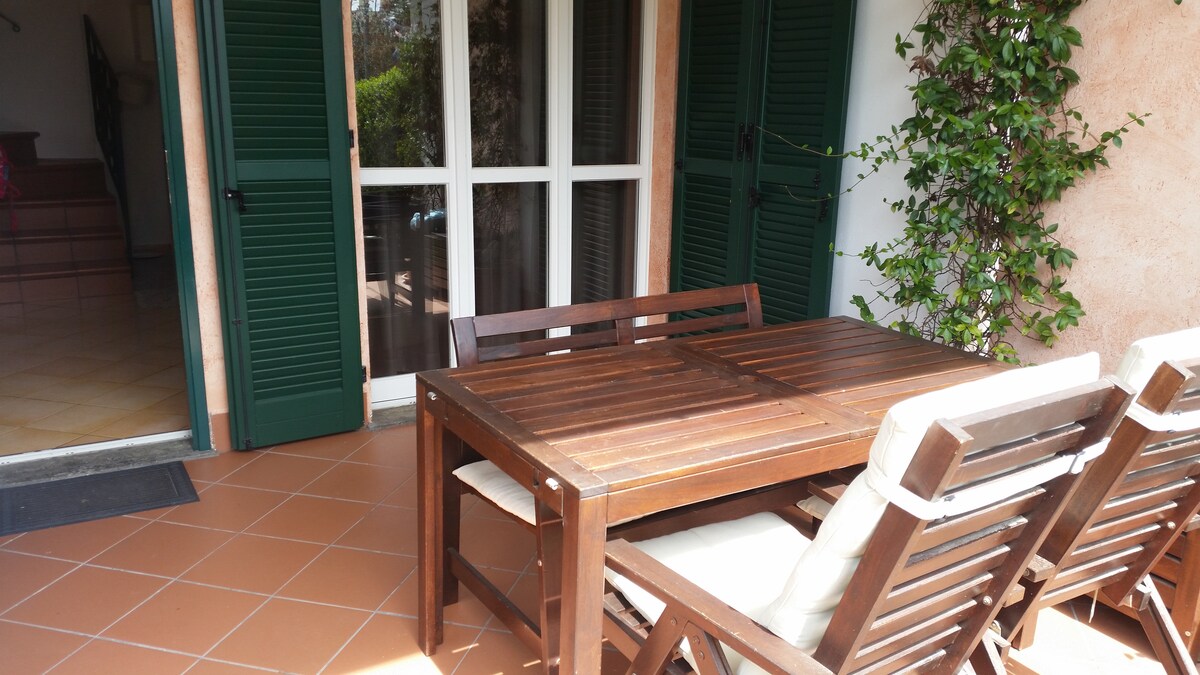 A spacious outdoor veranda is furnished with a wooden dining table surrounded by comfortable lounge chairs. Green shutters frame the entrance, and lush greenery is visible nearby, enhancing the inviting outdoor space suitable for dining or relaxation.