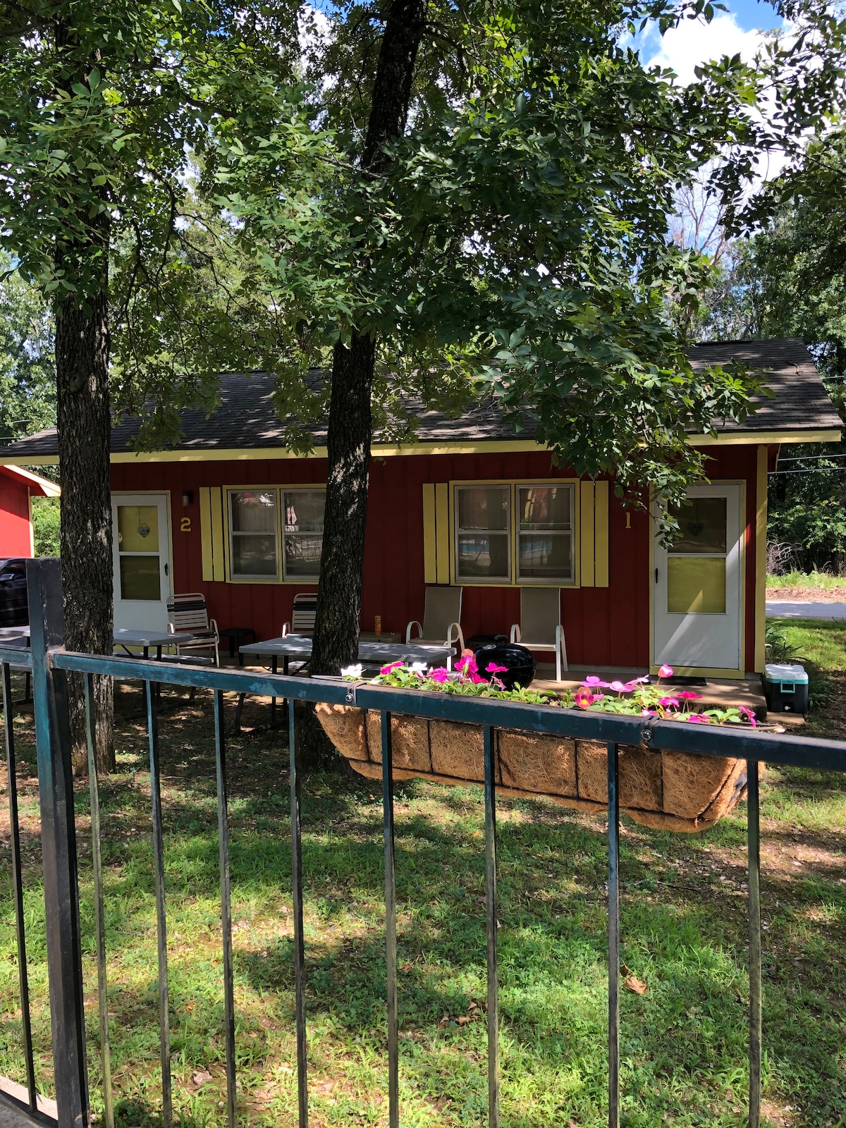 The exterior of the cottage is showcased, featuring a vibrant red facade with yellow shutters. Comfortable seating is visible under the shade of trees, complemented by colorful flowers in a hanging planter. The setting offers a peaceful view of the surrounding greenery.