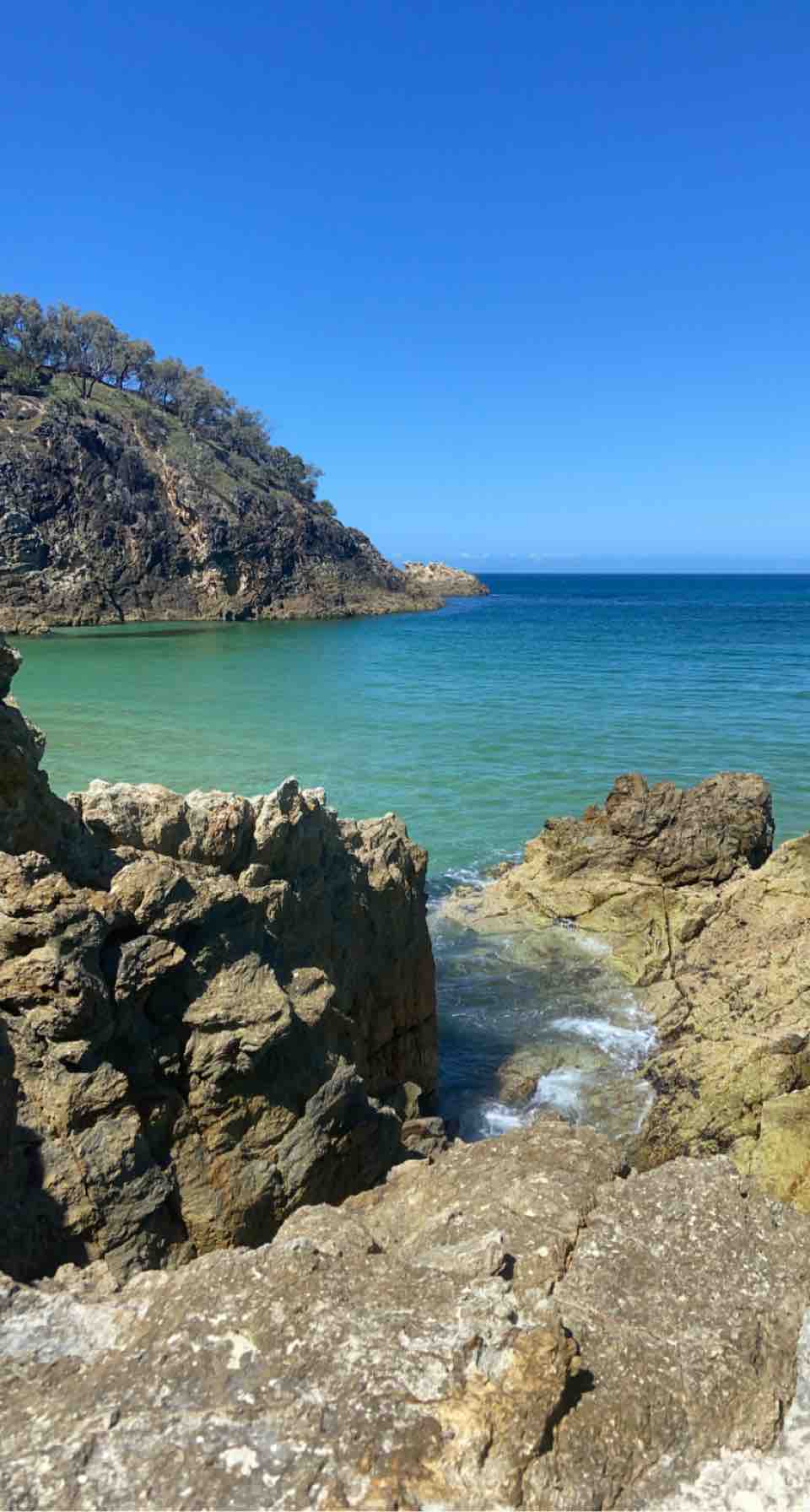 A rocky shoreline extends towards a calm, turquoise sea, with a distant landform rising in the background. The clear blue sky overhead reflects off the water, creating a serene coastal scene. Textures of the rugged rocks are prominent, contrasting with the gentle waves lapping at the shore.