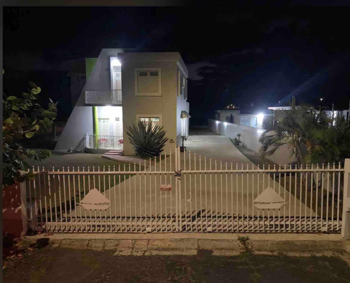A well-lit house is viewed from a distance at night, featuring a two-story design with a balcony and a large front yard. A gated entrance is visible, leading to a spacious driveway surrounded by palm trees and subtle landscape lighting.