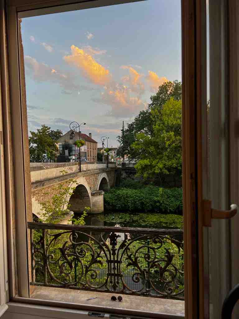 A scenic view from an open window displays a bridge arching over a gentle waterway, surrounded by lush greenery. Soft clouds are visible in the sky, reflecting warm hues as the day transitions to evening.