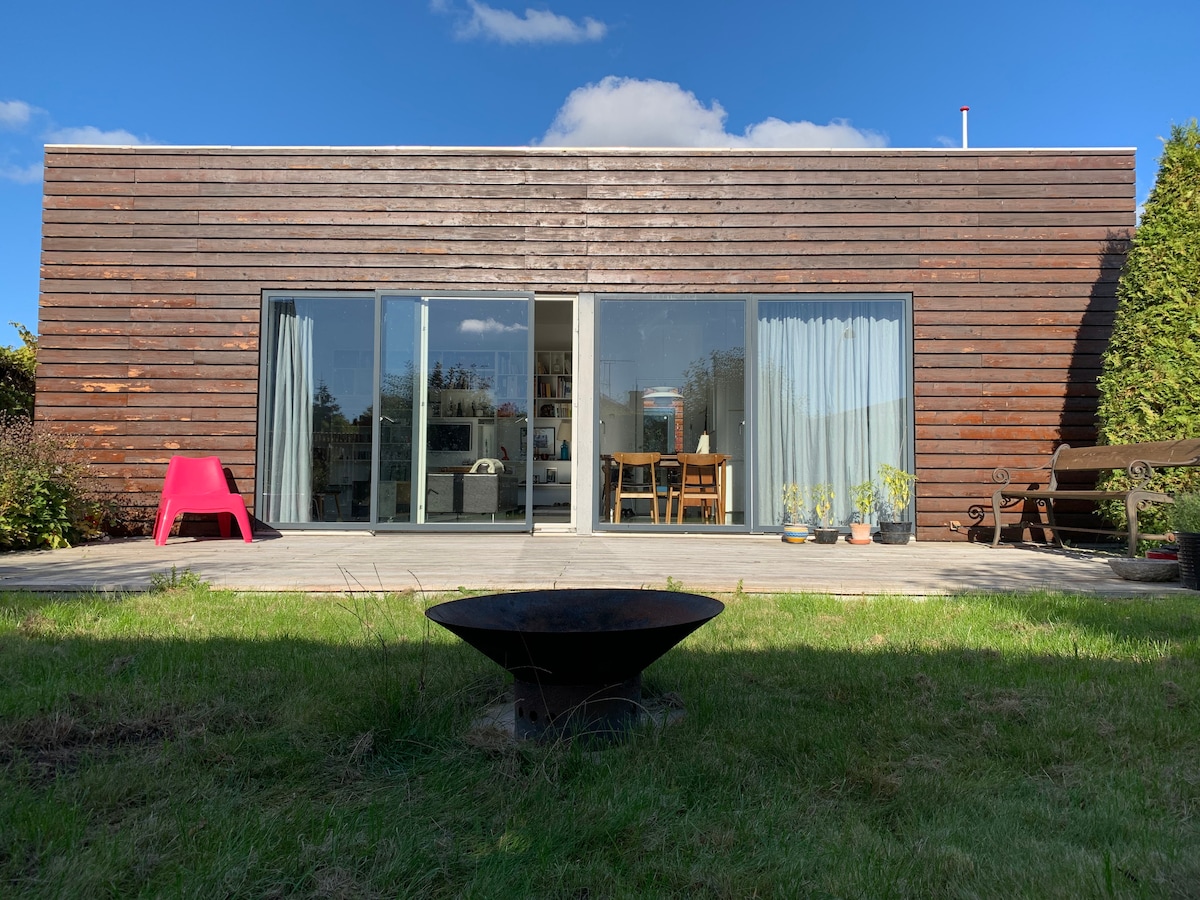 The exterior of a contemporary wooden house features large glass doors that open to a patio area. A table and chairs are visible inside, surrounded by greenery. A fire pit is positioned on the lawn, adding to the outdoor setting.