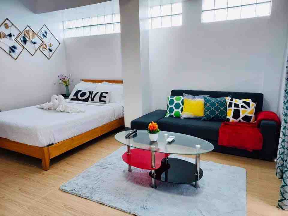 A bright studio apartment features a spacious bed adorned with a white blanket and decorative pillows. A black sofa, accented with colorful cushions, complements a glass coffee table situated on a soft area rug. Natural light filters through the high windows, creating an inviting space.