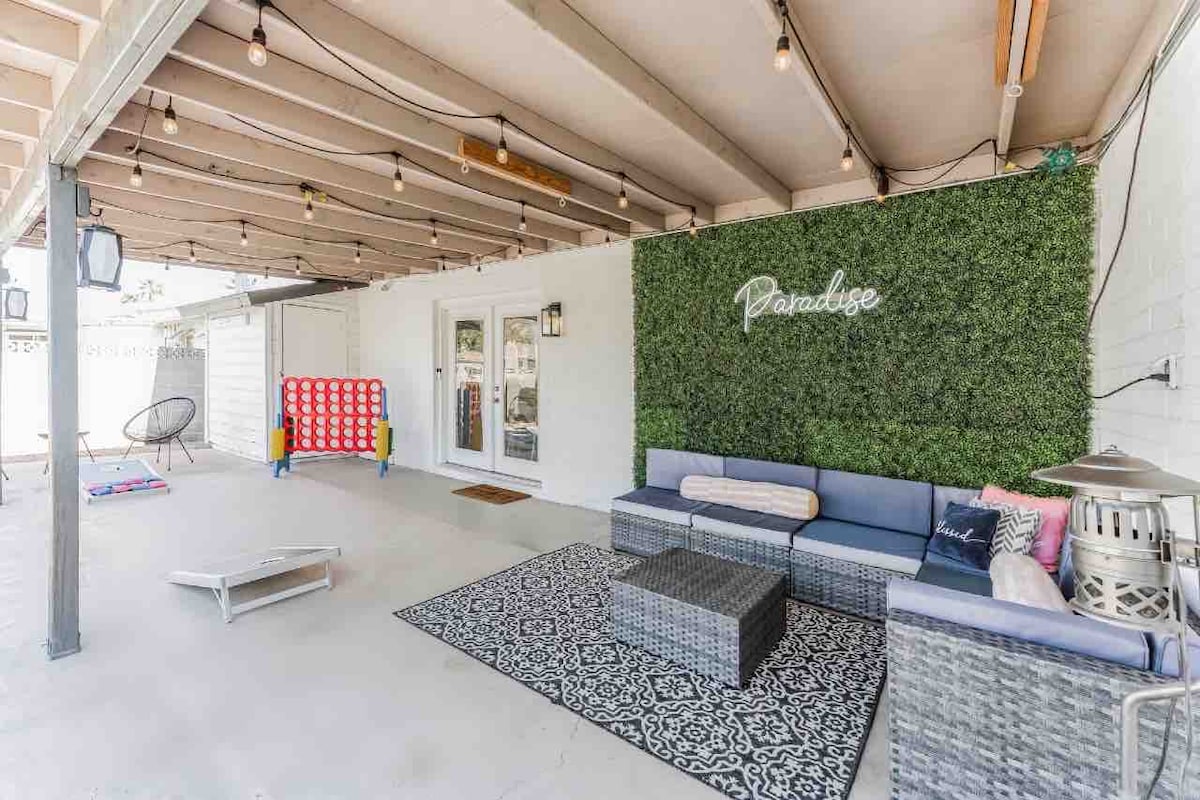 An inviting outdoor lounge area features a spacious sectional sofa, a patterned rug, and decorative wall greenery. String lights add ambiance overhead, while a children's game and individual seating options complete the space, providing comfort for gatherings.