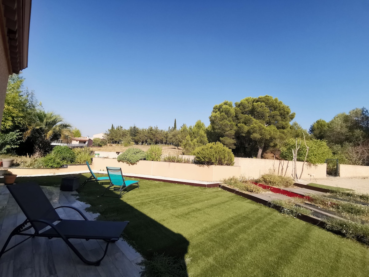 An outdoor space features a manicured lawn framed by lush greenery. Two lounge chairs are positioned on a stone patio, offering a view of the surrounding landscape. Tall trees and shrubbery create a serene atmosphere against a clear blue sky.