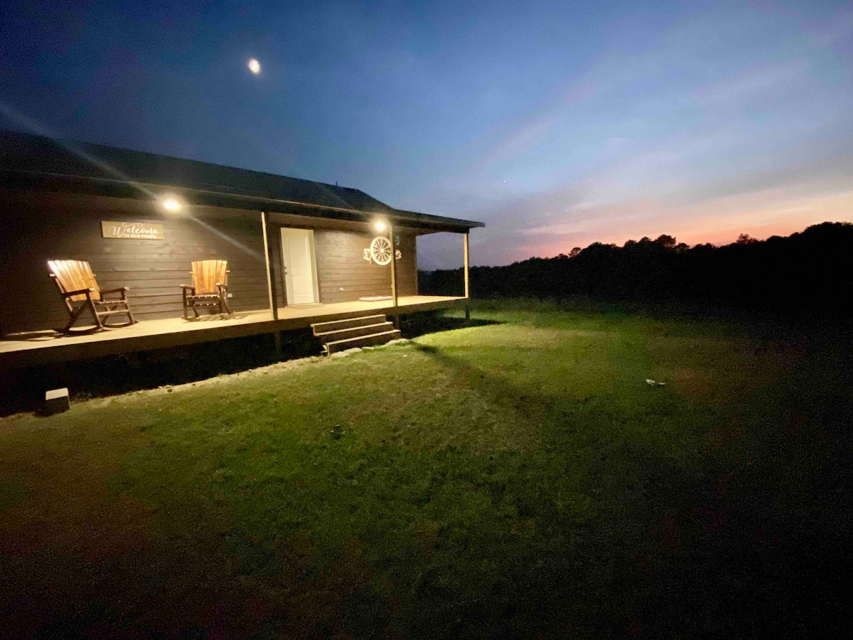 An isolated cabin is illuminated at dusk, highlighting two rocking chairs positioned on the deck. The expansive grassy area in the foreground gradually transitions to tree lines in the background, while a soft glow from the setting sun casts a serene ambiance.