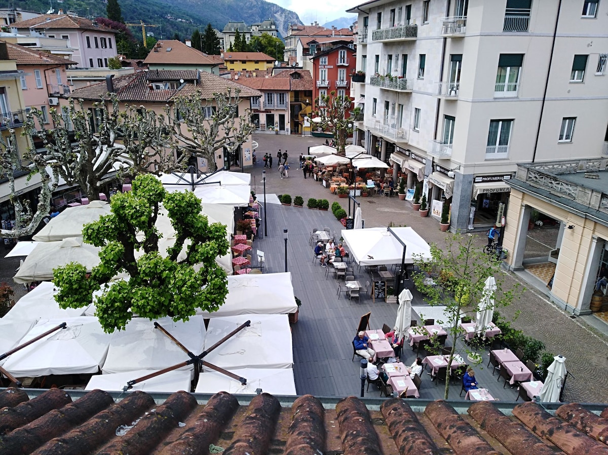 An aerial view of a lively piazza featuring outdoor dining areas adorned with umbrellas. Cafes and restaurants populate the space, with people gathered at tables. Lush trees provide greenery, and quaint buildings line the square, set against a backdrop of mountainous terrain.