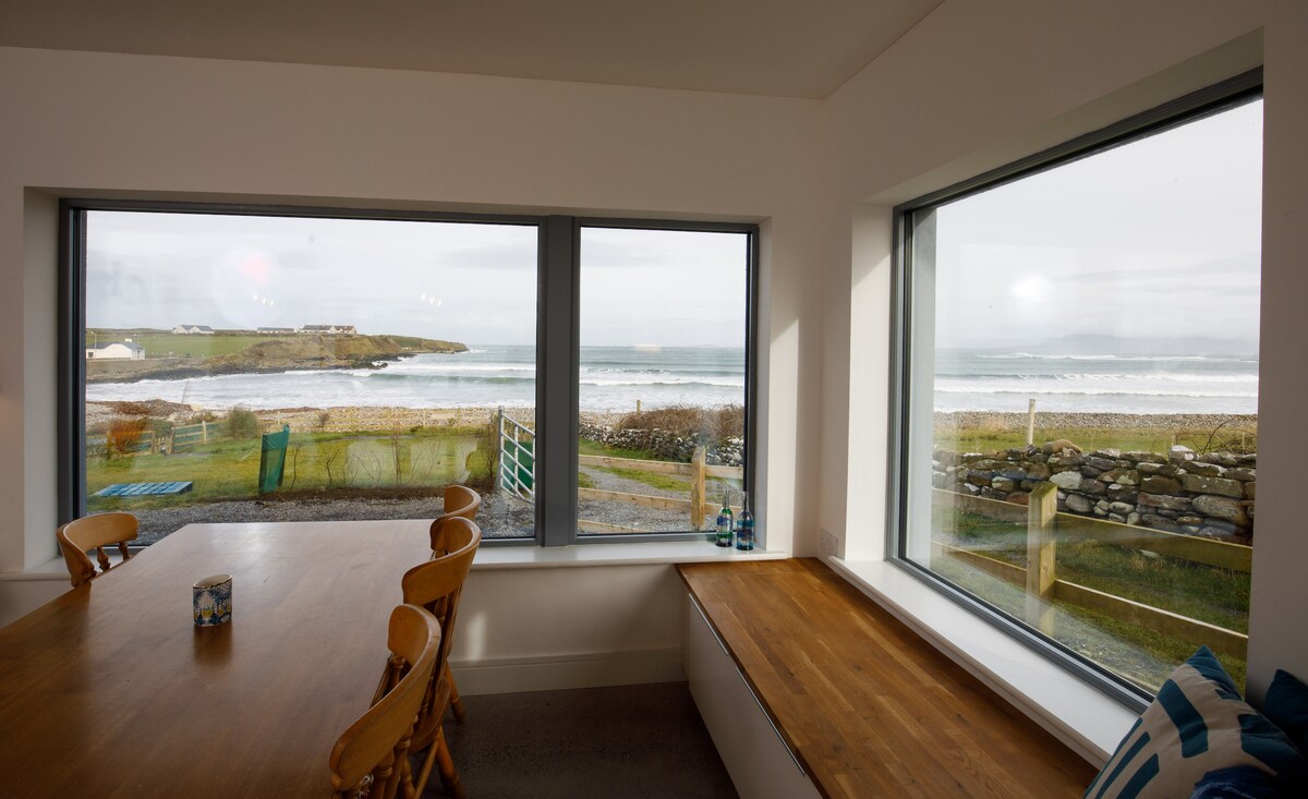 Large windows provide unobstructed views of the Atlantic Ocean and the surrounding landscape. A wooden dining table with six chairs is positioned within the open plan living space, enhancing the connection to the scenic outdoors. Natural light fills the room, creating a bright atmosphere.