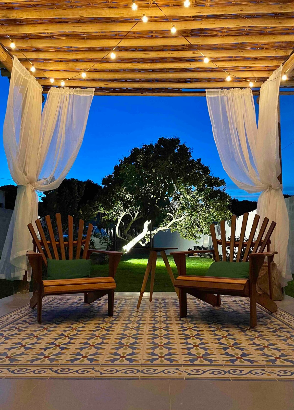 An outdoor seating area features two wooden chairs with green cushions, positioned around a small table. A decorative rug is laid beneath them. Soft lighting hangs from a pergola overhead, illuminating the space, while a nearby tree is subtly backlit in the evening atmosphere.