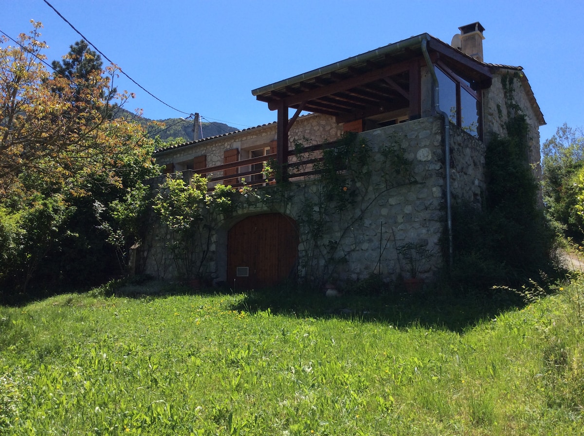 A charming stone house is nestled among greenery, featuring a balcony and large windows that invite natural light. The surrounding garden offers a spacious area, with trees providing shade. The picturesque hillside is visible in the background, enhancing the serene outdoor setting.