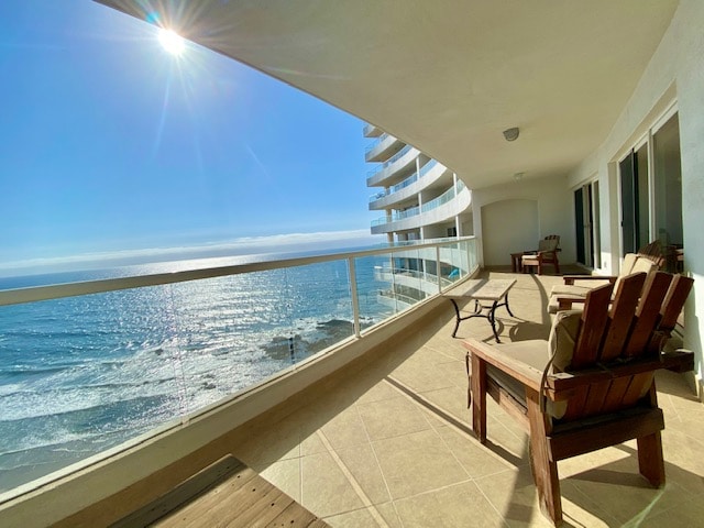 A spacious balcony features two wooden chairs and a small table, offering an unobstructed view of the ocean and coastline. The sun reflects off the water, creating a bright atmosphere. The tiled flooring complements the modern aesthetic of the building.