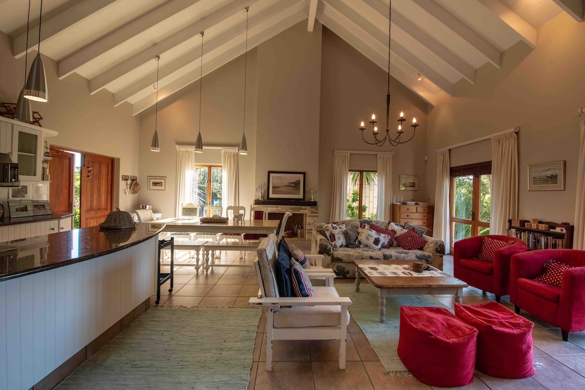 An open-plan living area features high ceilings with wooden beams and a large kitchen island. Comfortable seating options, including a sofa adorned with patterned cushions and red armchairs, invite relaxation. Natural light illuminates the room through large windows, enhancing the spacious atmosphere.