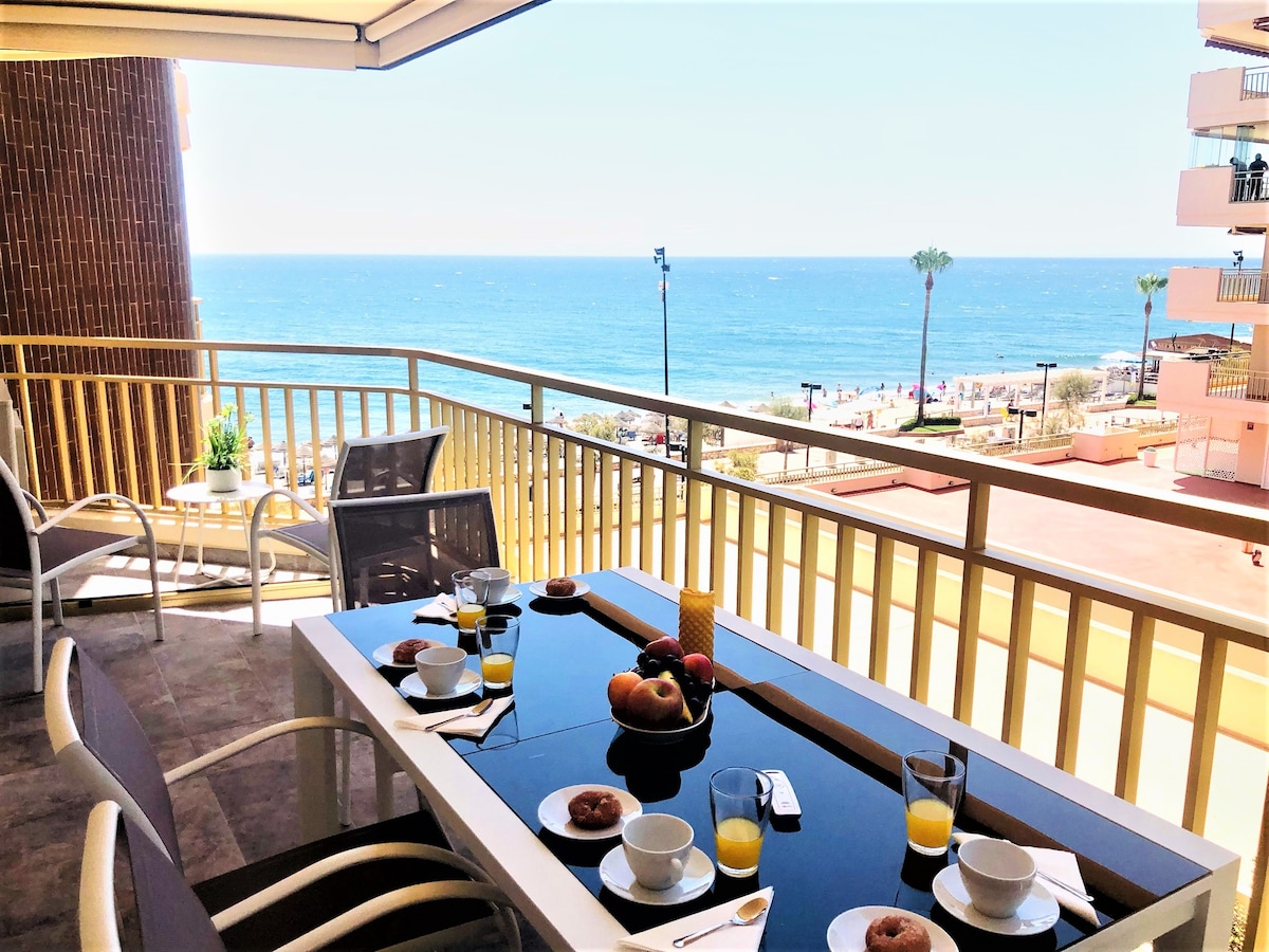 A spacious terrace is shown overlooking the beach, featuring a dining table set with plates, glasses, and a fruit centerpiece. Comfortable chairs are positioned around the table, while the ocean view extends in the background, complemented by clear blue skies.