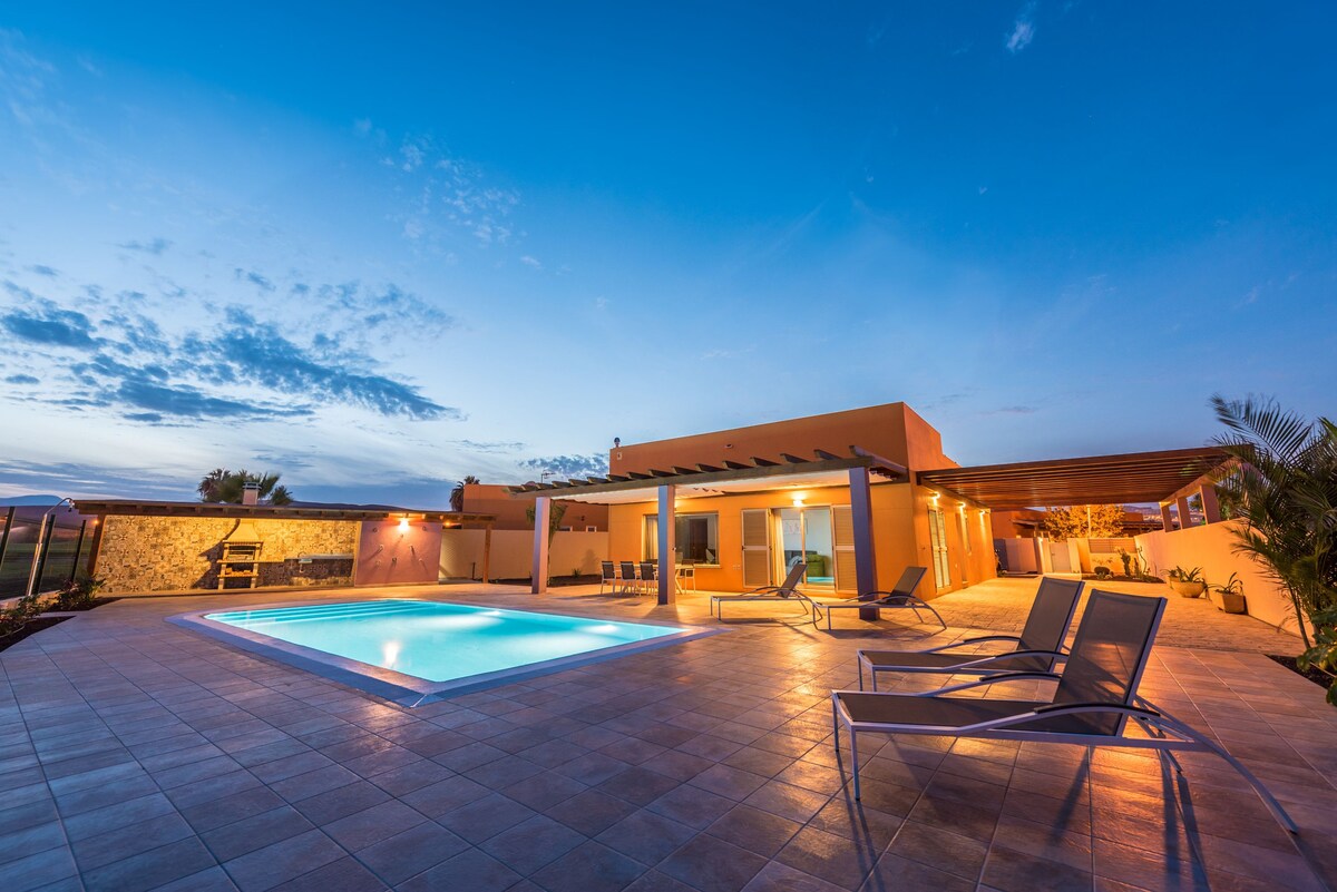 The outdoor area features a landscaped terrace with a heated swimming pool, surrounded by lounge chairs. Warm evening light enhances the modern architecture, while the sky transitions to twilight. A barbecue area is visible in the background, complementing the spacious layout.
