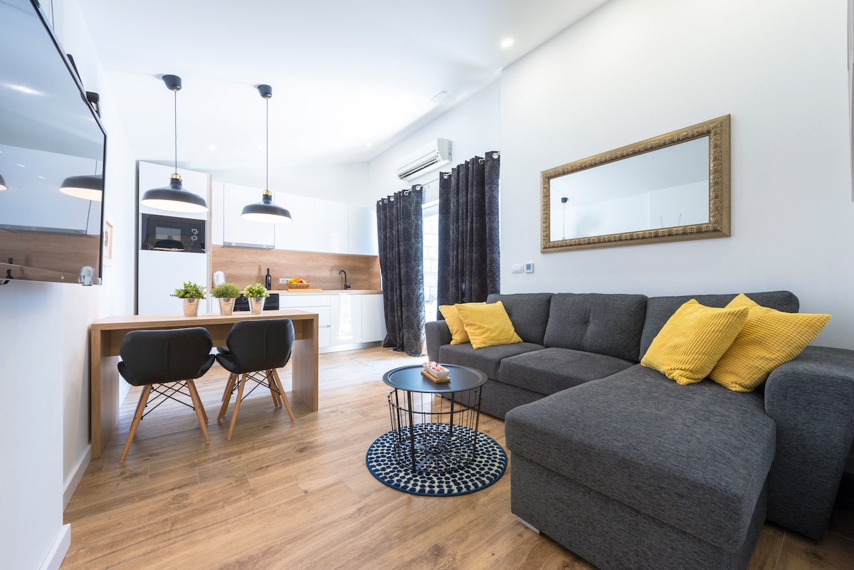 A modern living area features a spacious gray sofa with yellow accent pillows, paired with a round black coffee table. A wooden dining table with two chairs is positioned under stylish pendant lights. The room is complemented by a large mirror and patterned area rug.