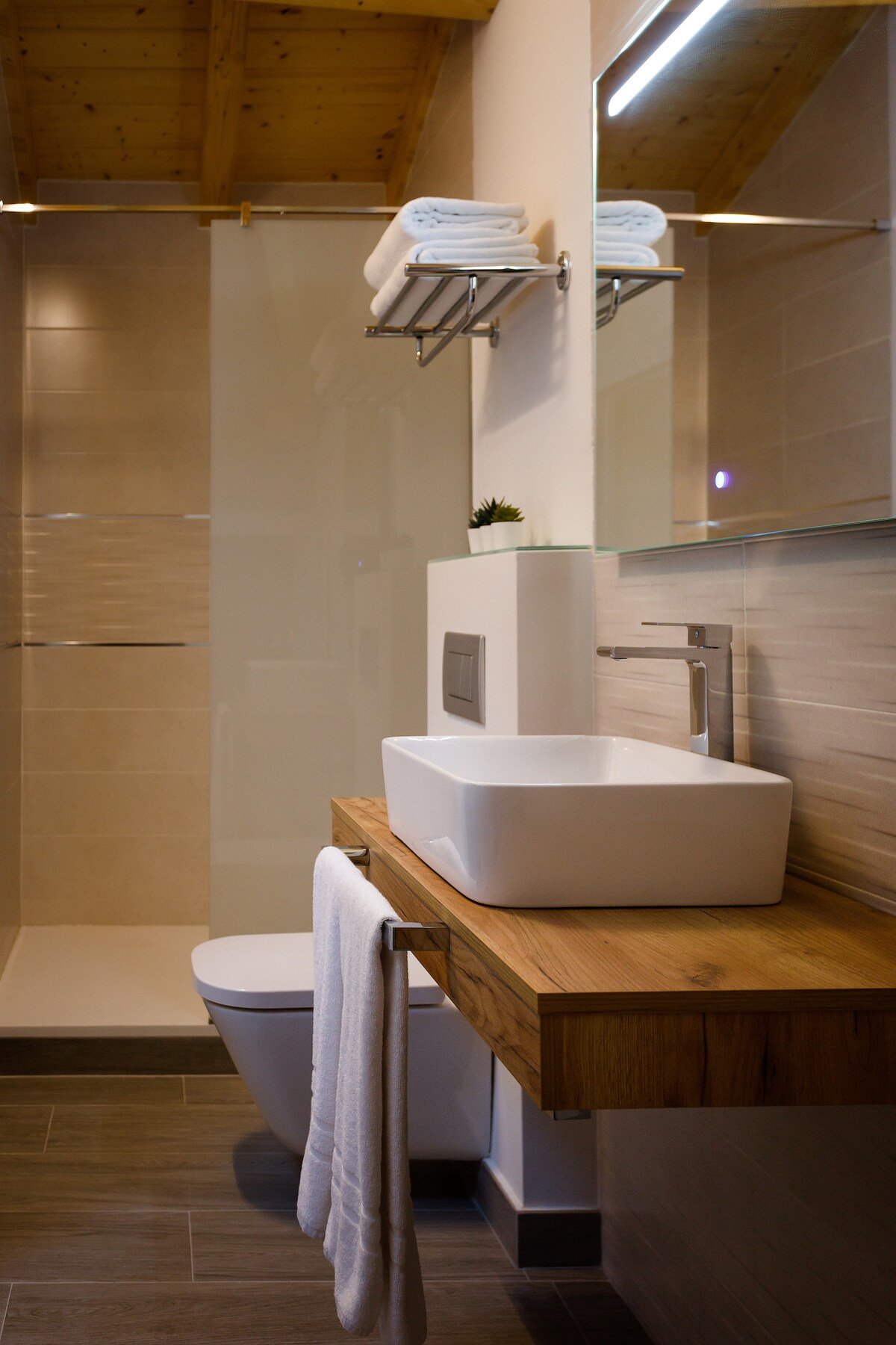 A modern bathroom is depicted, featuring a minimalist sink situated on a wooden countertop. Soft, ambient lighting illuminates the space, highlighting the stylish fixtures. A glass shower enclosure and neatly arranged towels on a shelf provide a sense of cleanliness and organization.