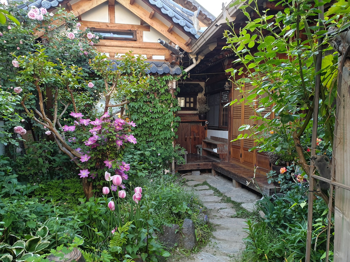 A serene courtyard surrounded by lush greenery features blooming pink roses and tulips. A stone pathway leads to the wooden structure with traditional Korean architecture, showcasing intricate roof details. The tranquil garden creates a peaceful atmosphere, inviting guests to relax among nature.