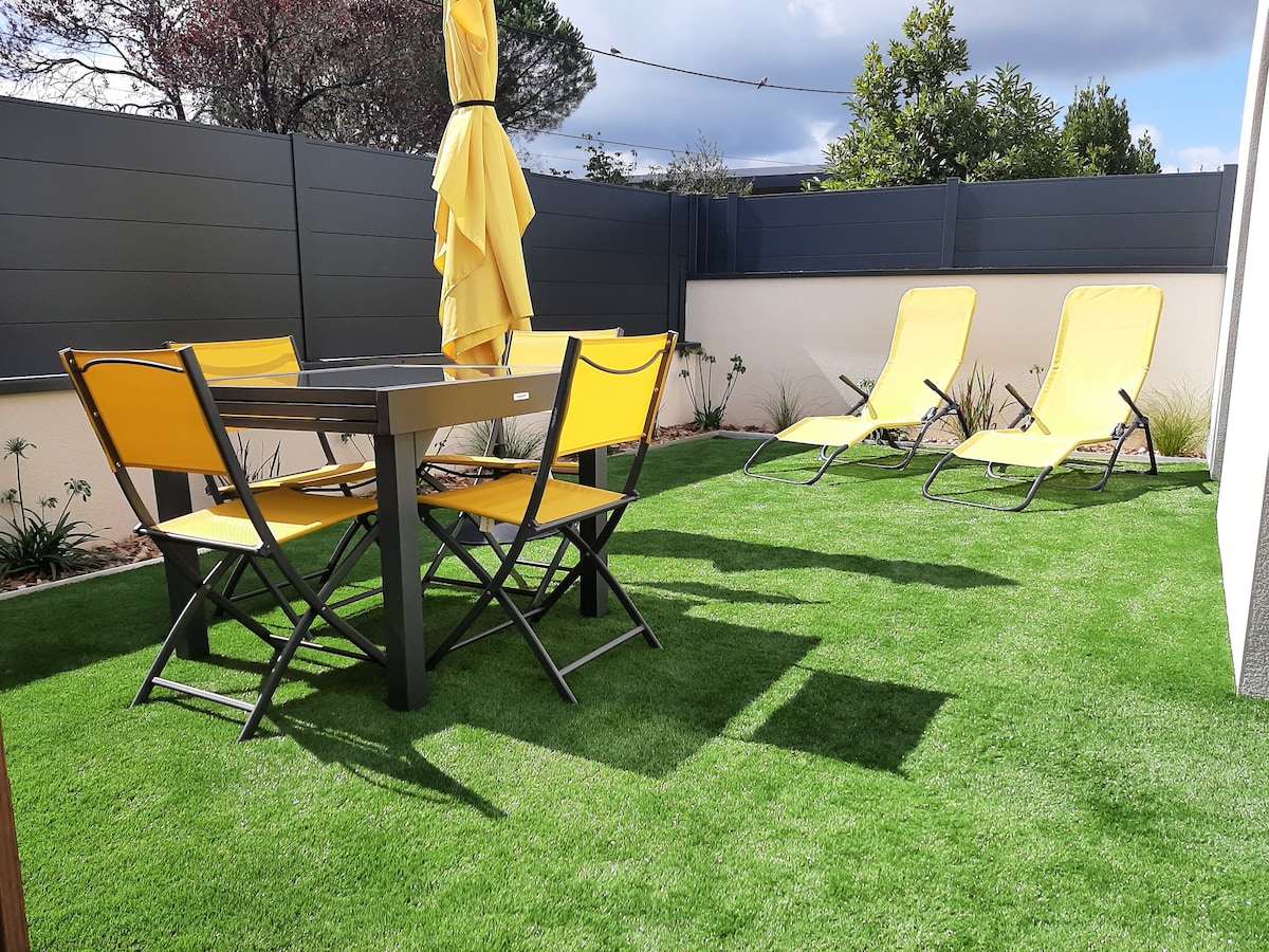 An outdoor area features a black table surrounded by four yellow folding chairs and two reclining lounge chairs. A yellow umbrella stands at the table, providing shade. Lush green artificial grass enhances the space, bordered by well-kept landscaping and a privacy fence.