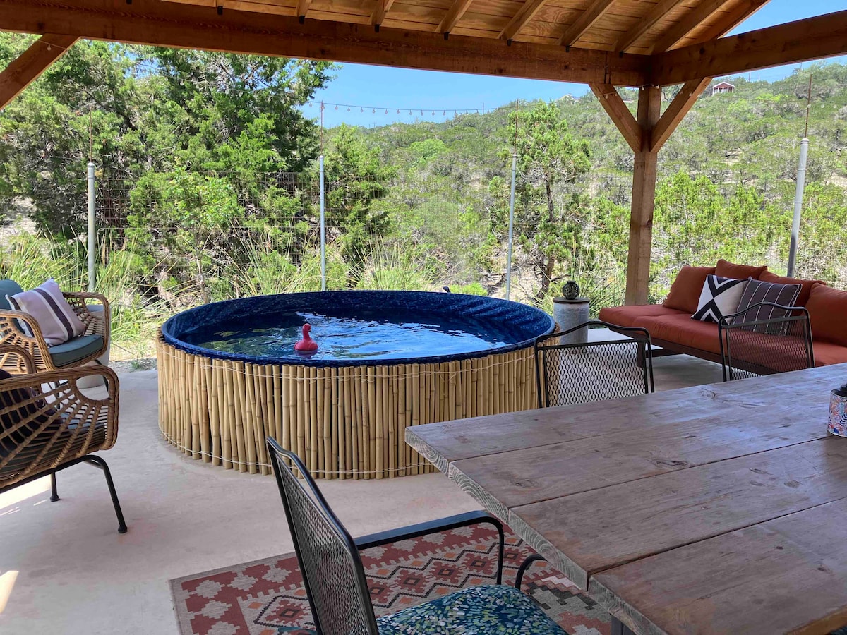 A stock tank pool is surrounded by natural scenery, featuring a vibrant blue interior. Nearby, a wooden dining table is positioned under a pavilion, with comfortable seating and decorative pillows. The area is enhanced by greenery, creating a serene atmosphere for relaxation.
