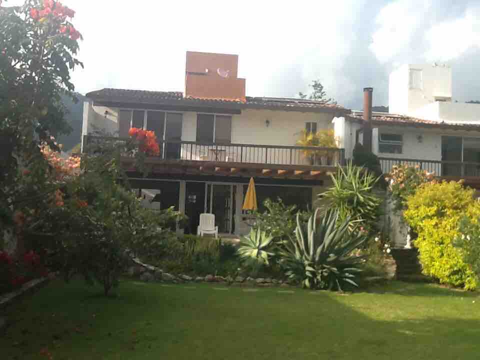 The image shows a two-story house surrounded by a lush garden, featuring a wooden terrace with a yellow umbrella. Colorful flowers and greenery add vibrancy, while a stone pathway leads through the garden, enhancing the tranquil outdoor setting.