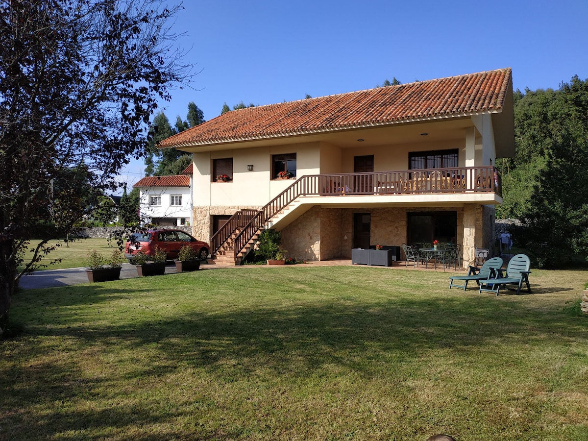 A two-story chalet is set in a spacious garden, marked by green grass and trees. The upper balcony overlooks the yard, featuring a wooden railing. Outdoor seating is arranged on the ground level, with a vehicle parked nearby. Clear blue skies complement the scene.