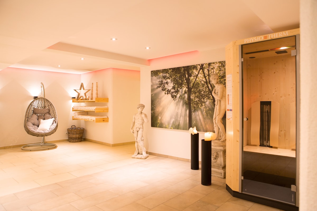 The wellness area features a serene atmosphere with soft lighting. A hanging chair provides a cozy seating option, while decorative sculptures are positioned throughout the space. A wooden sauna blends harmoniously with the environment, and a large artwork depicting a sunlit forest adds a tranquil touch.