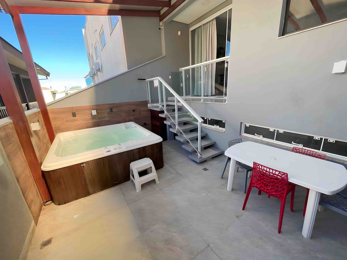 A private outdoor space features a heated spa and a dining area. Several steps lead to the spa, surrounded by a wooden enclosure. A white table with colorful chairs accommodates dining or relaxation, complemented by the sleek stone flooring and clean, modern design.