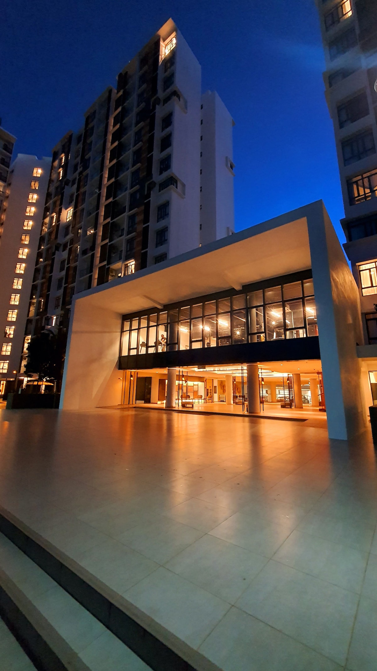 An elegantly designed building is depicted during dusk, featuring a large glass facade that reflects the surrounding lights. The spacious outdoor area is paved with tiles, and the setting conveys a modern architectural style, inviting engagement with the space.
