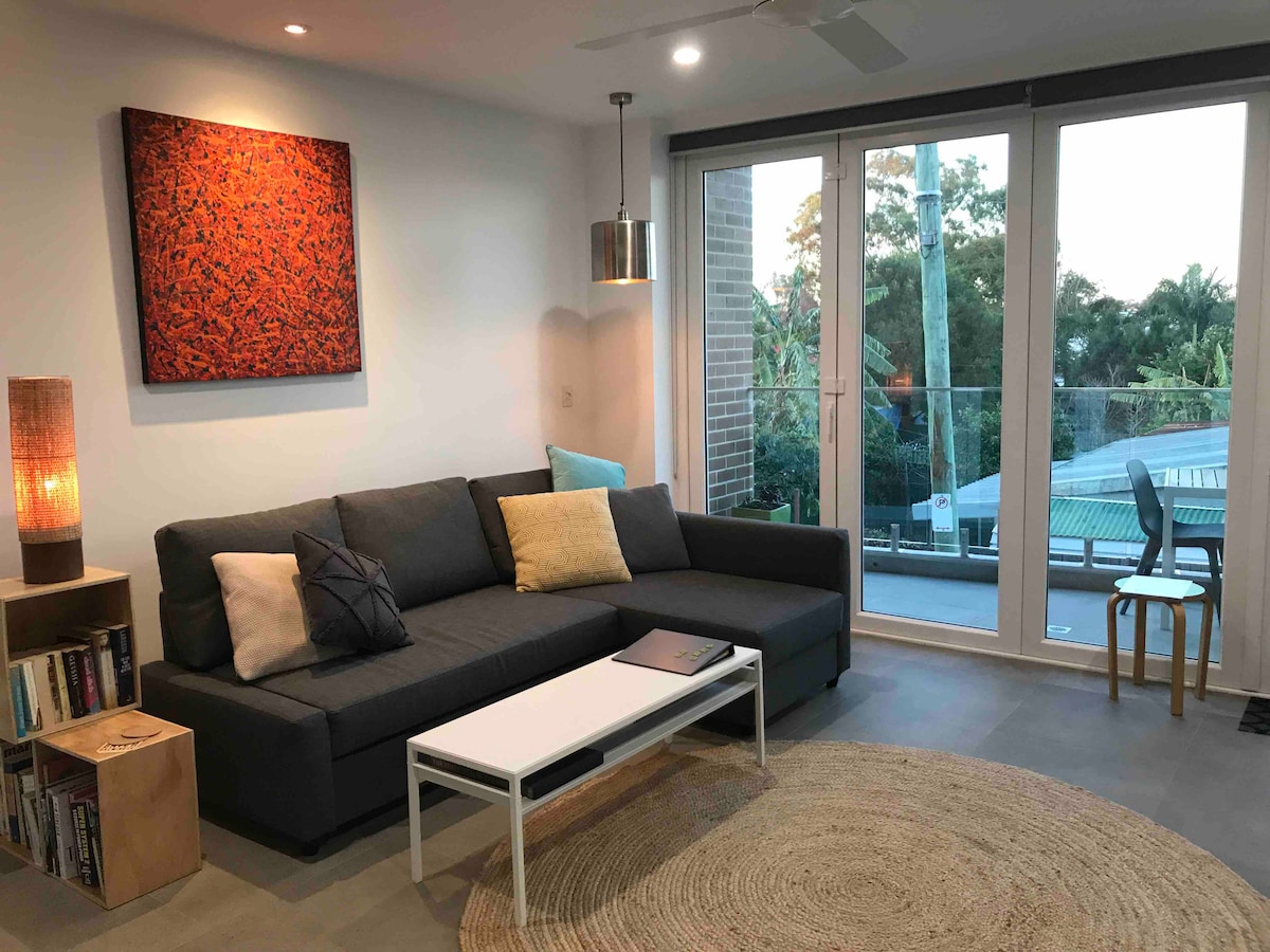 A modern living area features a spacious gray sectional sofa adorned with decorative cushions. A white coffee table is positioned in front, while a striking piece of artwork hangs above. Floor-to-ceiling sliding doors lead to a balcony, allowing natural light to fill the room.