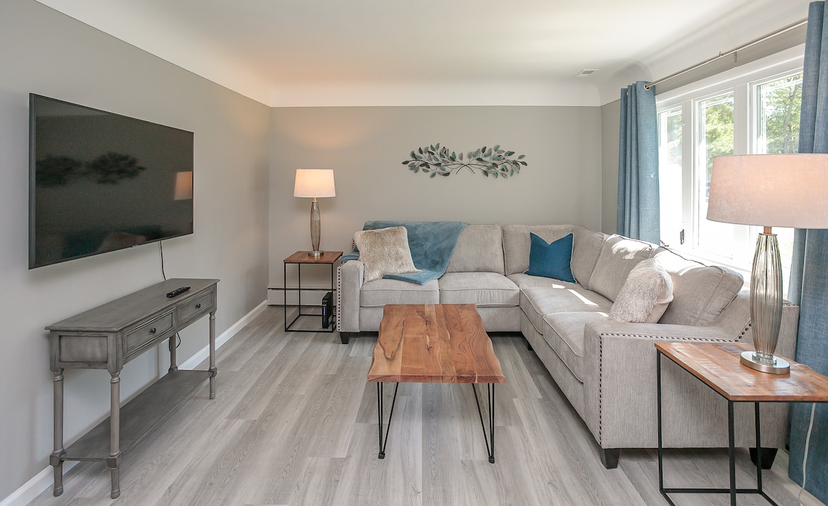 A living room is showcased, featuring a sectional sofa with light gray upholstery and decorative pillows. A wooden coffee table sits center, complemented by side tables. A flat-screen TV is mounted on the wall opposite a large window, allowing natural light to fill the space.