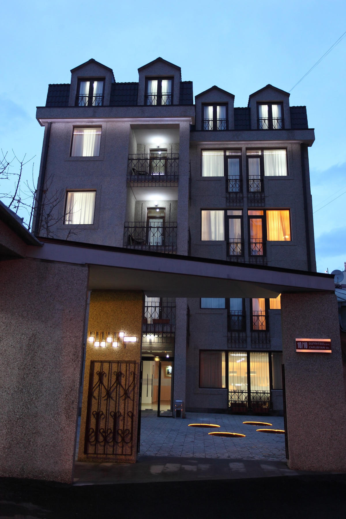 The exterior of the Cascade Hotel is captured at dusk, showcasing a four-story building with a classic architectural style. Illuminated windows reflect a warm glow, while ornamental balcony railings add character. The entrance features an inviting pathway with subtly lit design elements underfoot.