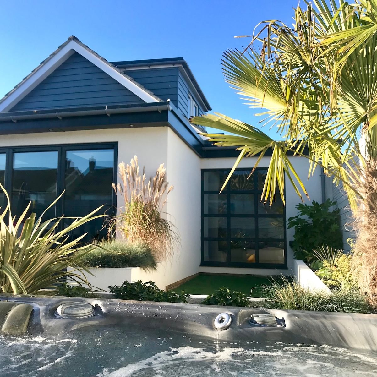 The exterior of a modern house is visible, framed by lush greenery, including palm trees and ornamental grasses. Large windows reflect sunlight, enhancing the contemporary design. A corner of a hot tub is shown in the foreground, inviting relaxation amidst the vibrant surroundings.