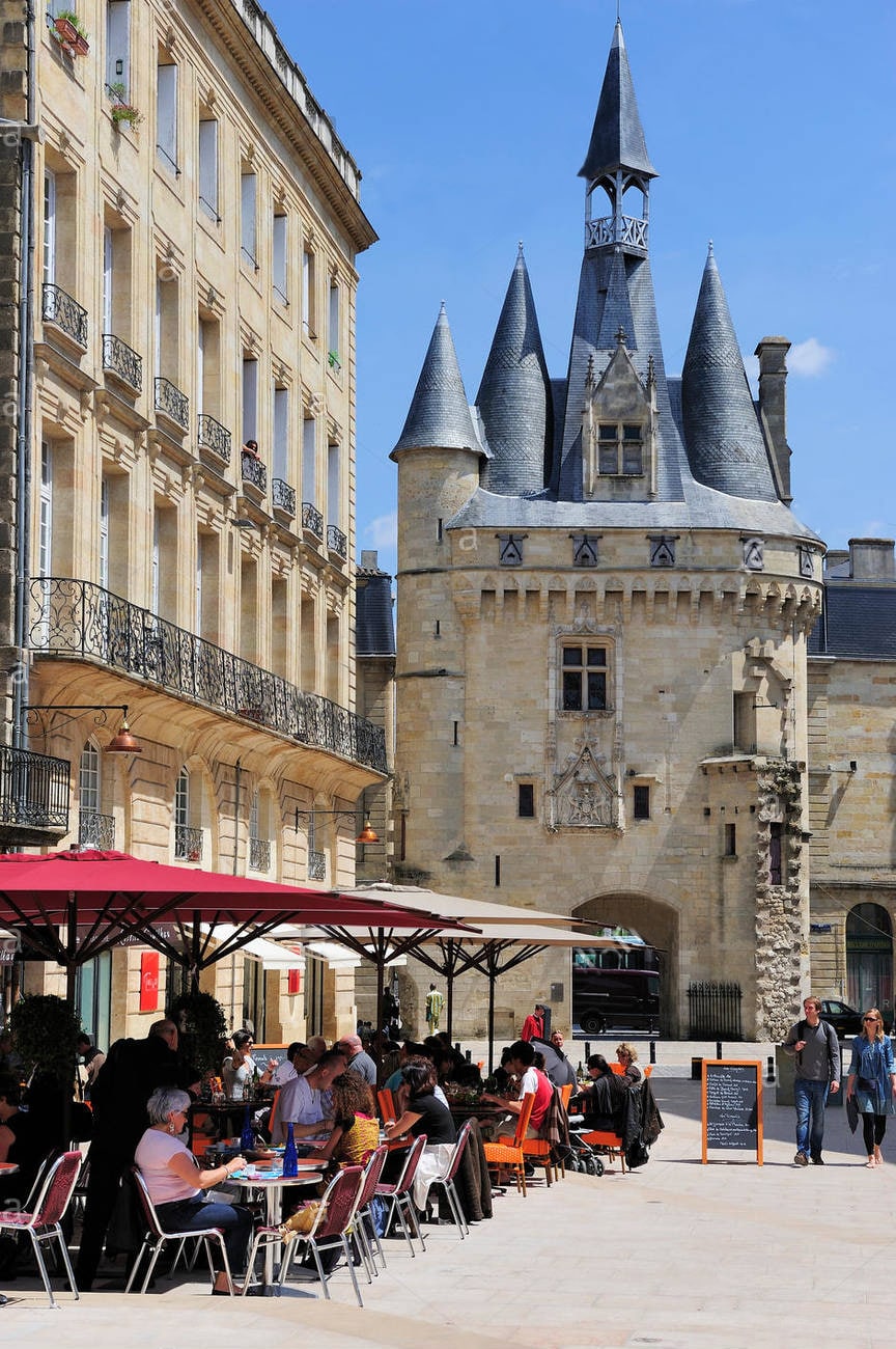 A lively outdoor dining area is visible, with several tables covered by red umbrellas. Patrons are present, enjoying their meals. In the background, a historic stone building features distinct spires and architectural details, adding character to the vibrant street scene.